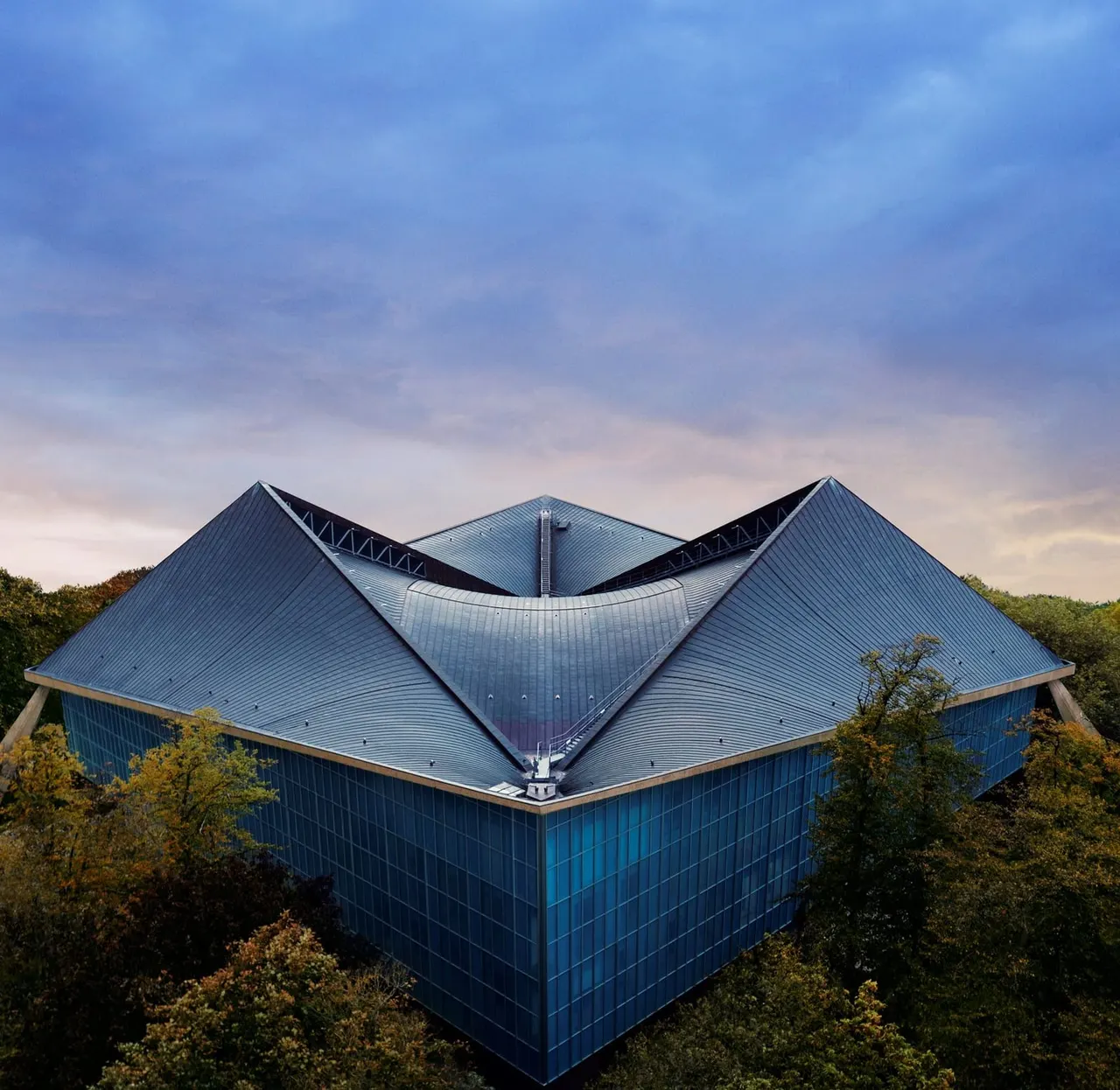Modern blue building with distinctive angular roof set amongst autumn woodland under cloudy sky.