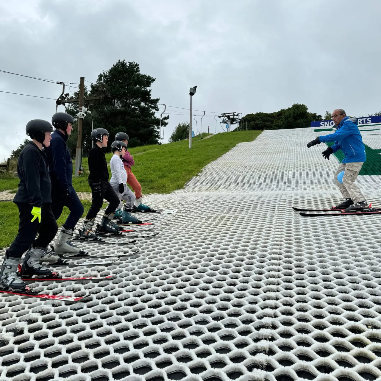 Ski instructor demonstrating technique to a group of skiers wearing helmets on an indoor dry slope.