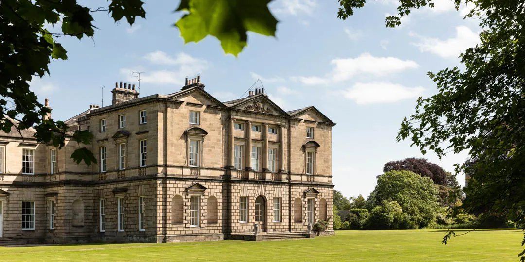 Grand stone mansion set in manicured grounds, framed by green foliage and blue sky.