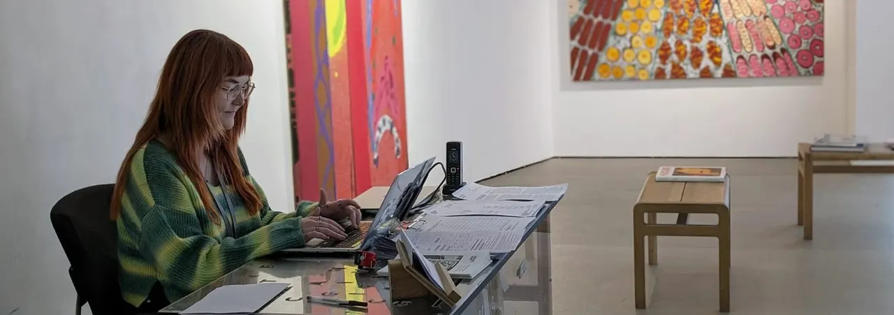 Woman working at desk with laptop in art gallery surrounded by colourful paintings.