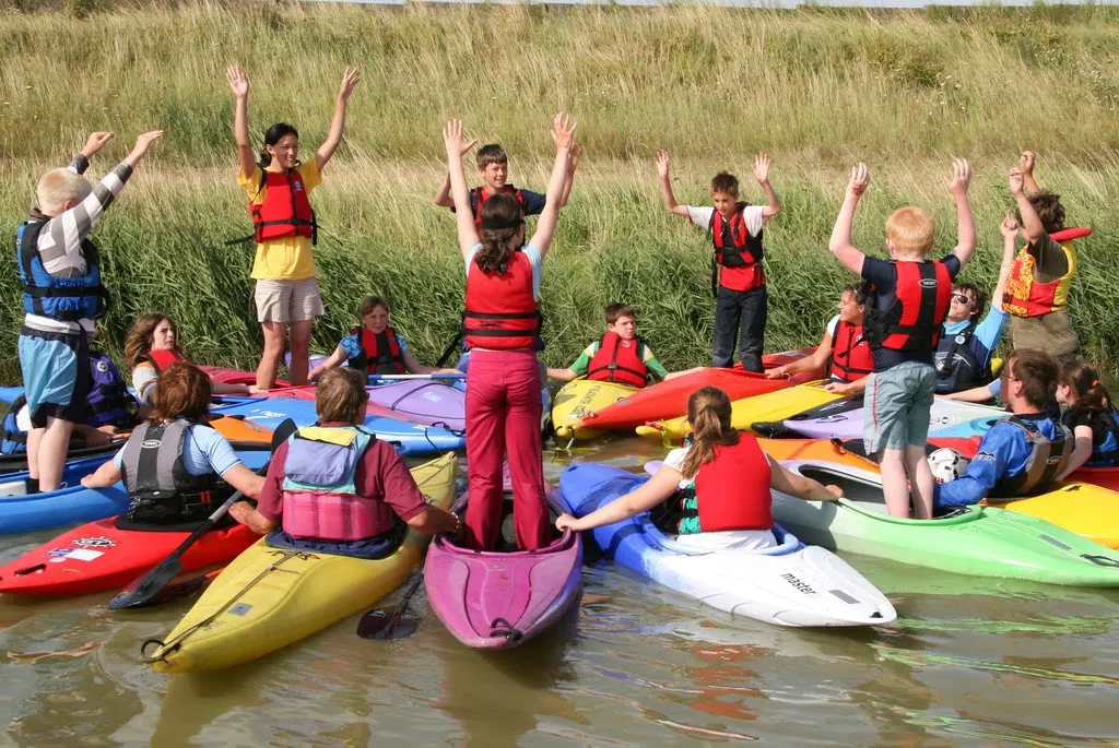 Children in life jackets celebrating at a water's edge with colourful kayaks during a water sports activity.