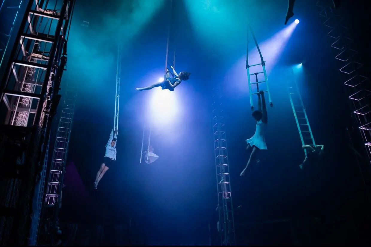Acrobats perform on suspended ladders and silks against blue lighting during a circus performance.