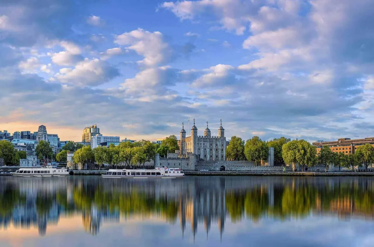 Historic fortress with golden spires reflected in calm river water, with passenger boats and London skyline beyond.