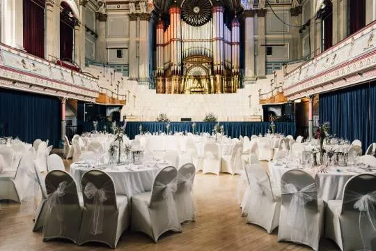 Elegant event space with round tables dressed in white linens and grey chair covers, ornate organ visible at far end of grand hall.