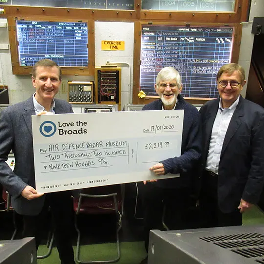 Three men in business attire holding a large cheque for a charity donation inside a museum control room with vintage radar equipment.