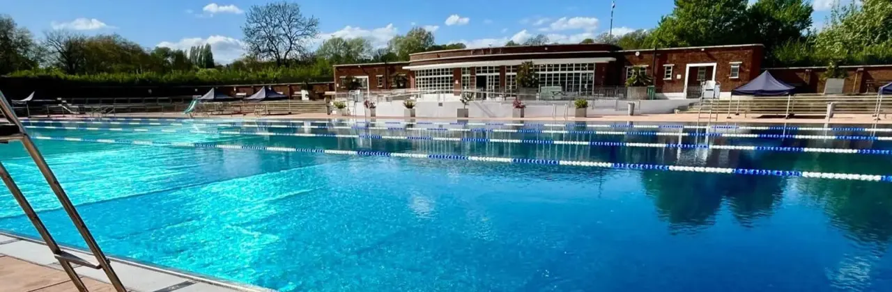 An outdoor swimming pool with lane dividers, surrounded by a brick building, trees, and clear blue sky.