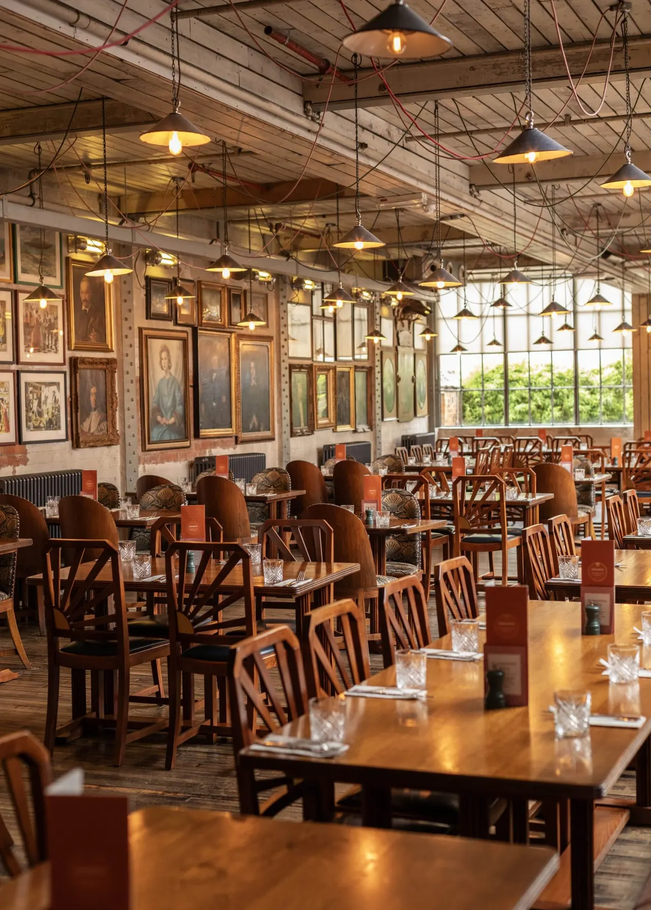 Industrial-style restaurant interior with wooden tables, upholstered chairs, pendant lights and framed artwork on walls.