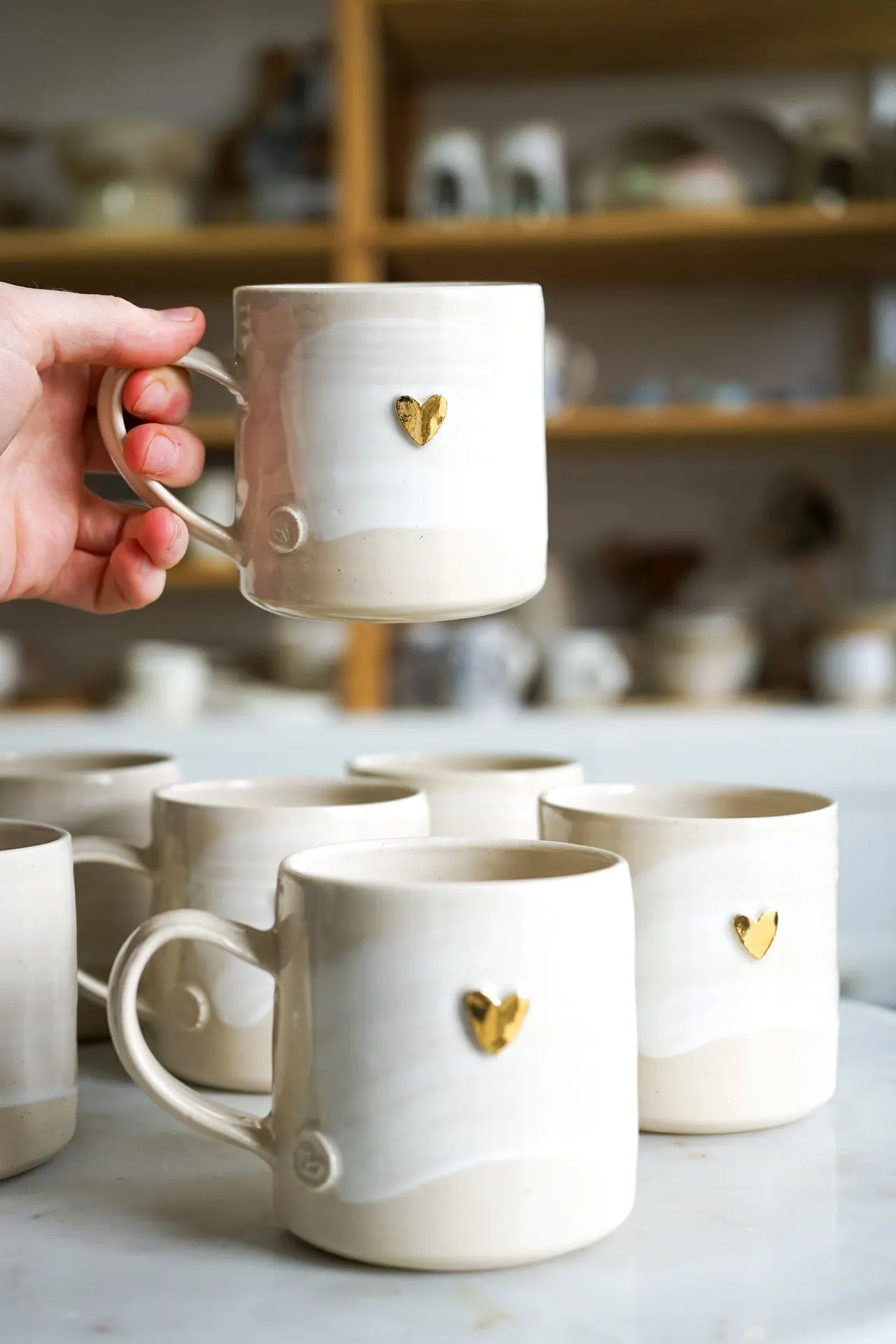 A hand holding a cream ceramic mug decorated with a small gold heart, displayed among similar mugs in a pottery studio.