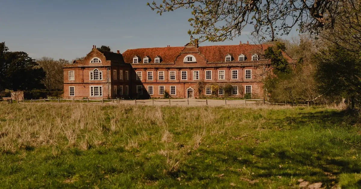 Large red-brick Georgian manor house with mansard roof set behind open parkland and fence.
