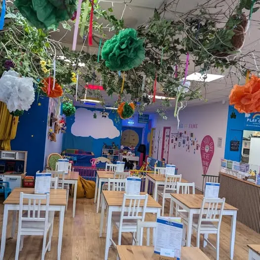 An indoor play café with wooden tables and white chairs decorated with hanging paper flowers and foliage from the ceiling.