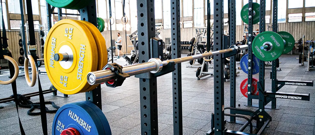 Interior of a functional fitness gym with multiple weightlifting racks loaded with coloured plates and barbells, people training in the background.