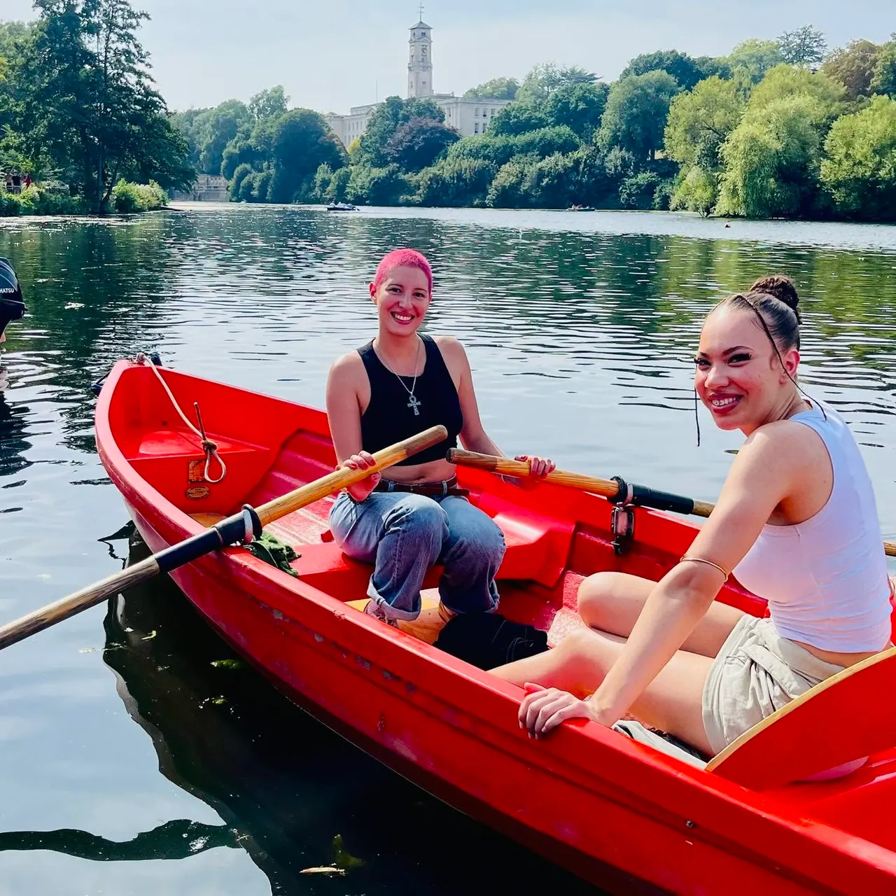 Two young women kayaking in a red boat on a calm lake, with trees and a historic church tower visible across the water.