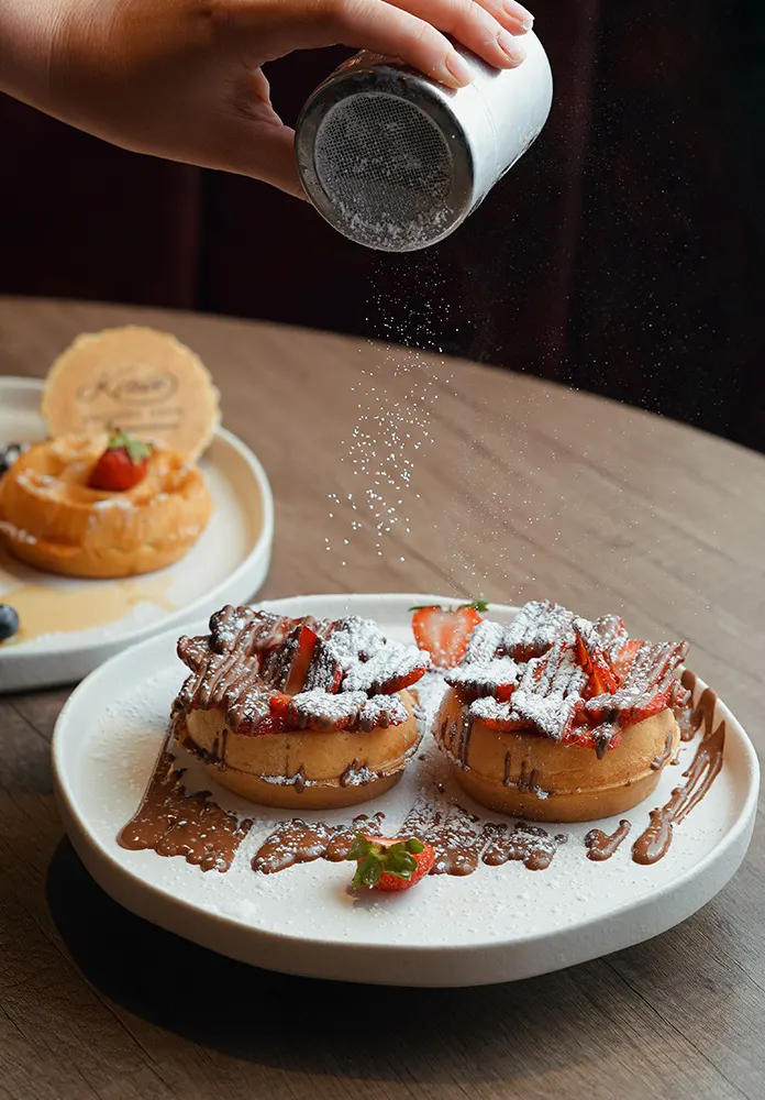 Hand dusting icing sugar over chocolate pastries topped with strawberries on a white plate.