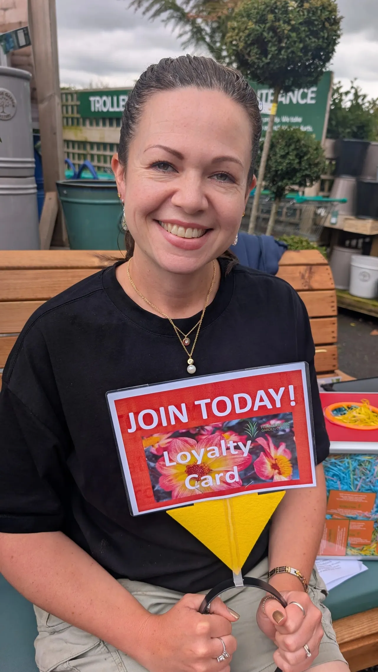 Staff member smiling at outdoor garden centre holding loyalty card promotion sign.