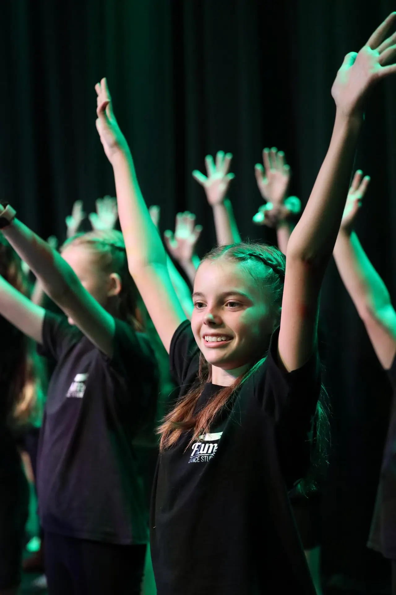 Young dancers in black outfits performing on stage with raised arms under green lighting.
