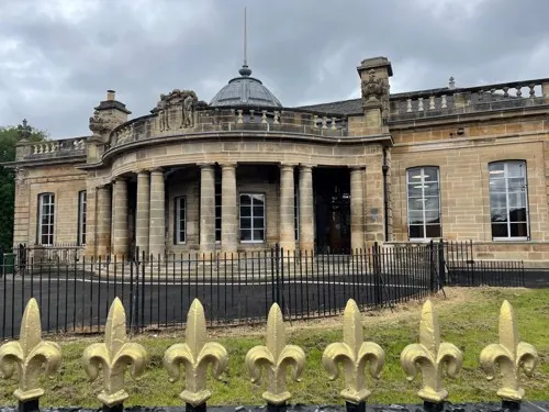 Stone neoclassical building with columned portico and domed roof, surrounded by ornamental fence.