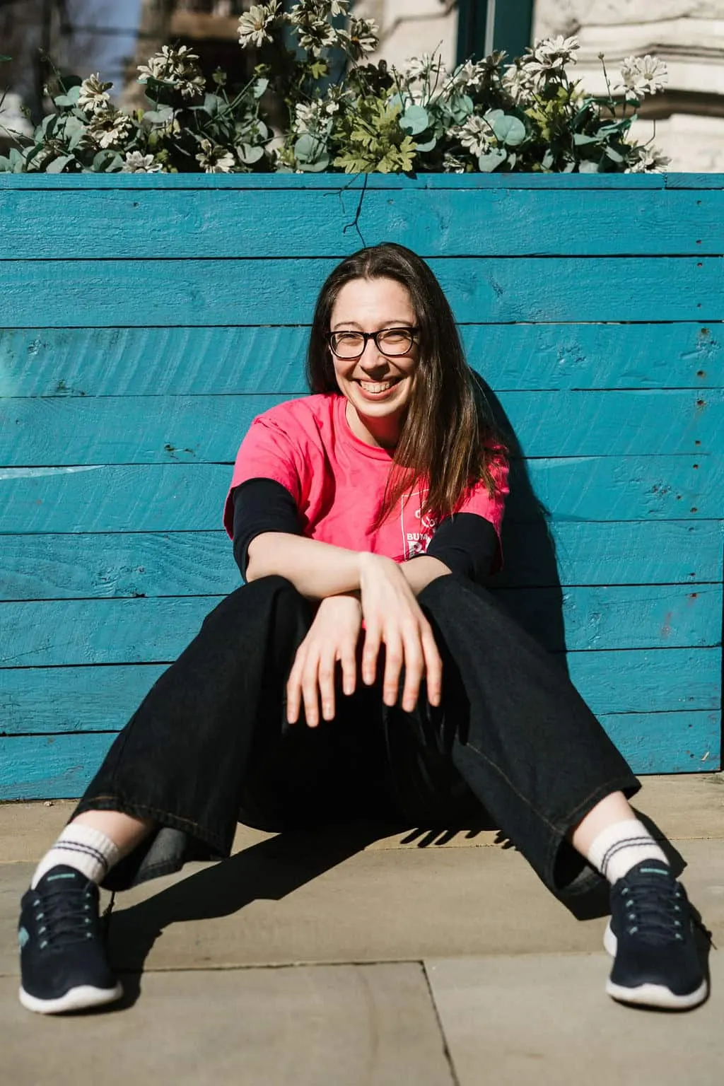 Young person sitting cross-legged in front of a bright turquoise wooden wall with white flowers above.