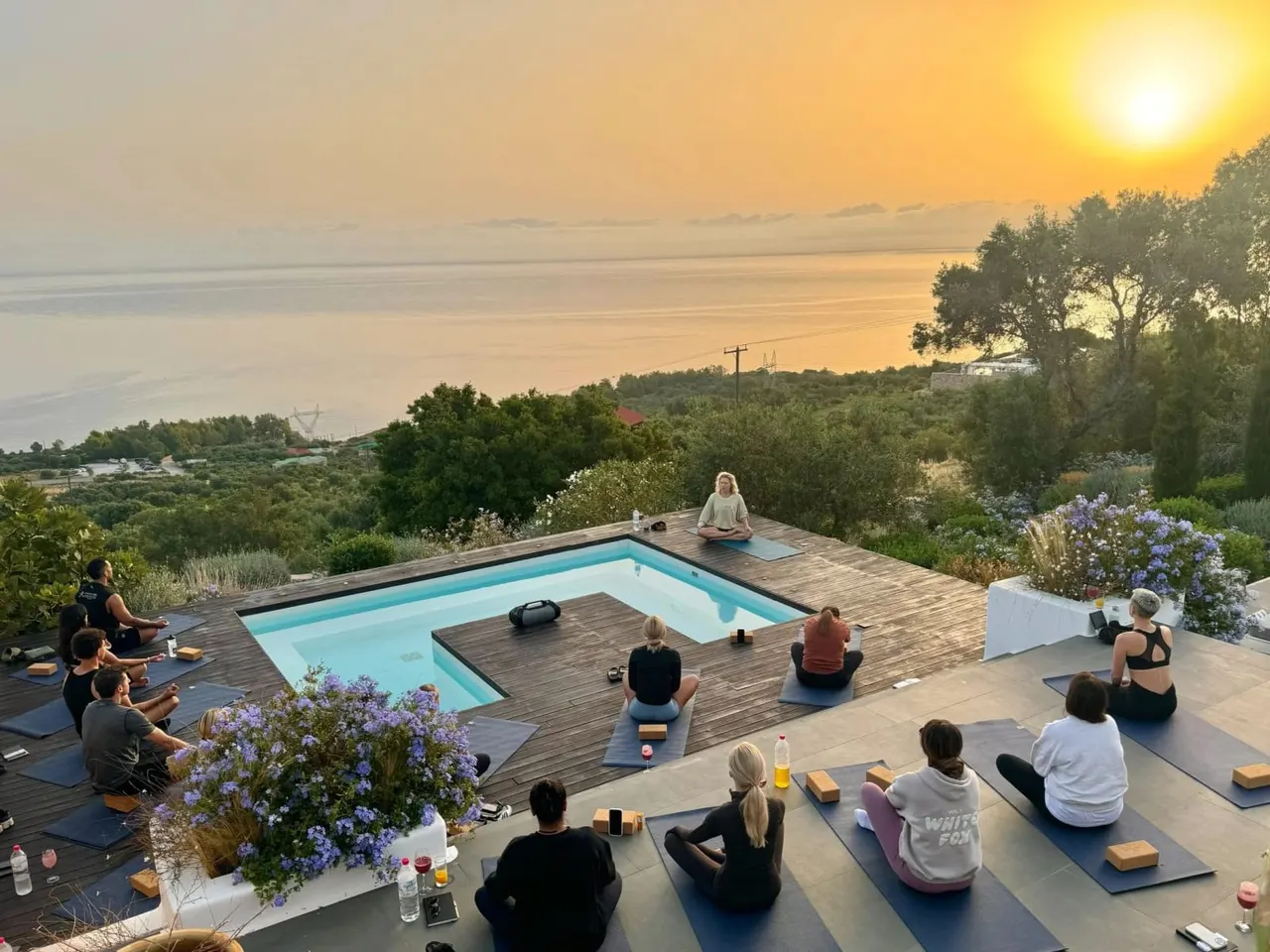 Group of people practising yoga on mats around an infinity pool overlooking a lake at sunset.