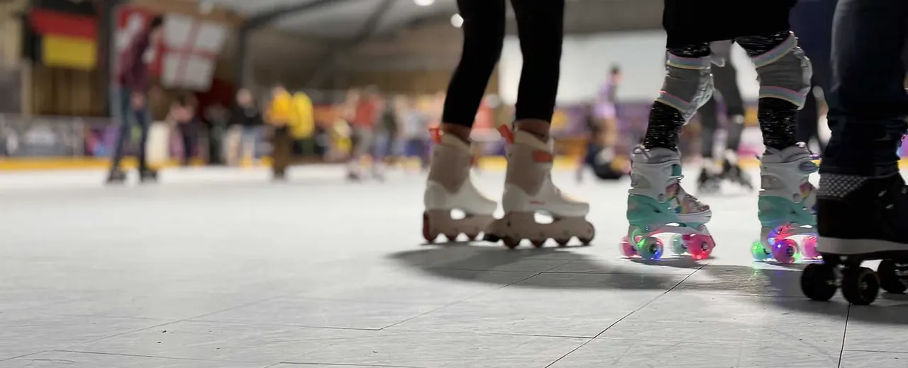 Children wearing roller skates and quad skates on an indoor skating rink floor with spectators in the background.
