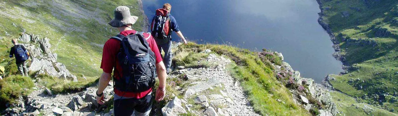 Hikers with backpacks walking along a rocky mountain ridge with steep grassy slopes and a lake visible in the valley below.