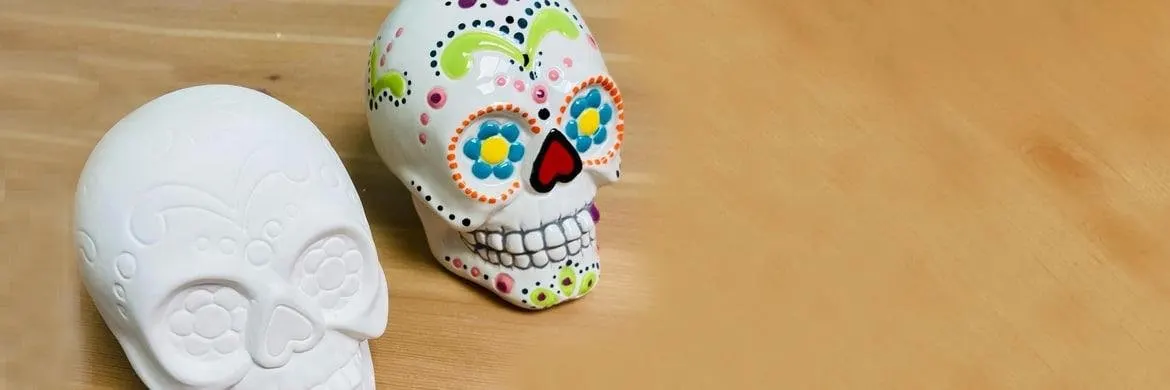 Two ceramic skull ornaments on a wooden surface, one plain white and one hand-painted with colourful flowers and decorative details.