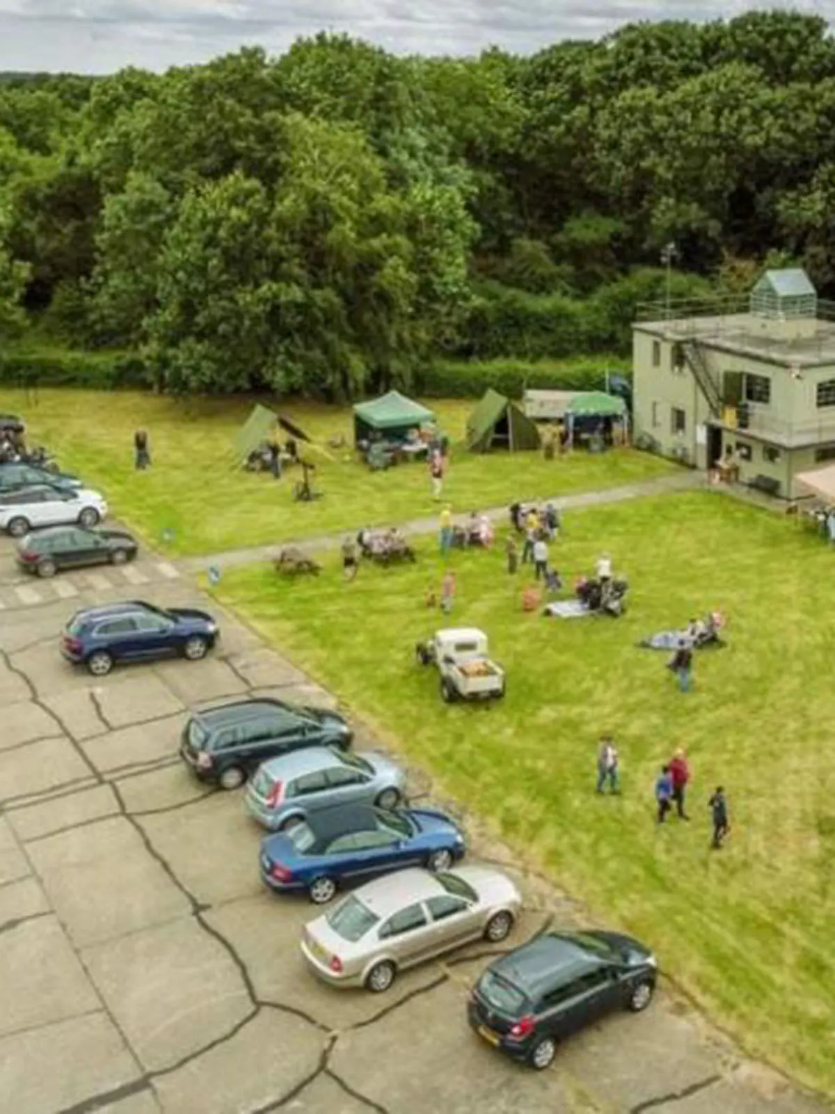 Aerial view of a grassy field with visitors, parked cars, a building, and woodland beyond.