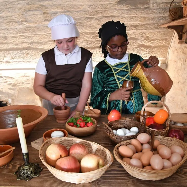 Two children in historical costume preparing food with fresh produce and eggs at a wooden table indoors.
