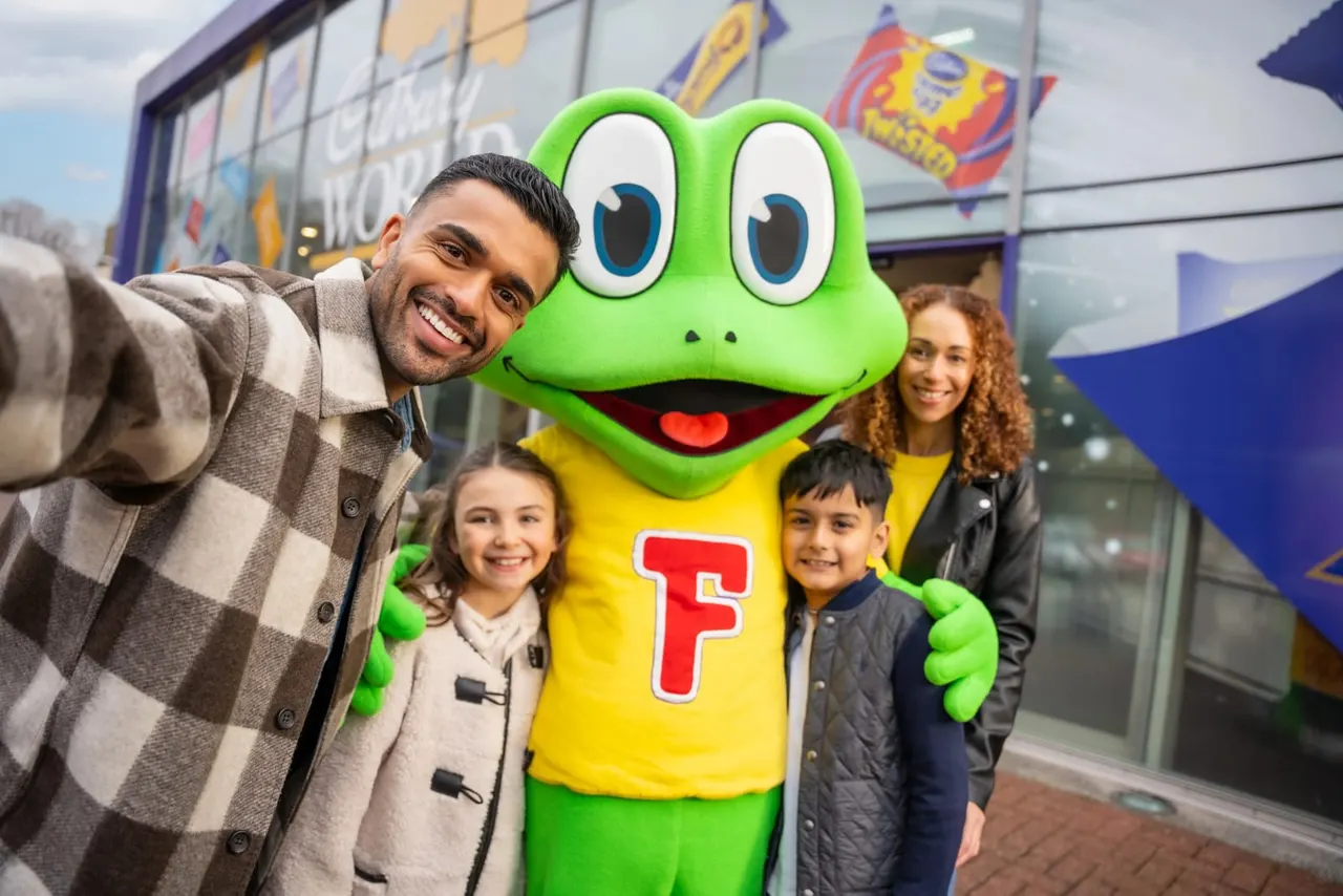 Family posing with a green frog mascot character wearing a yellow shirt outside Cadbury World.