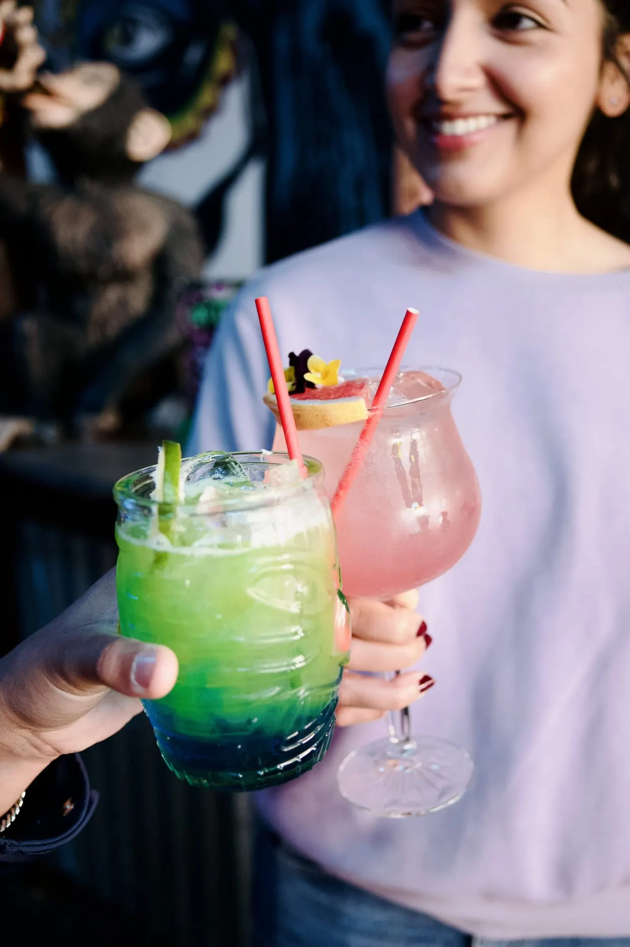 Woman holding two colourful cocktails with straws and fruit garnishes outdoors.