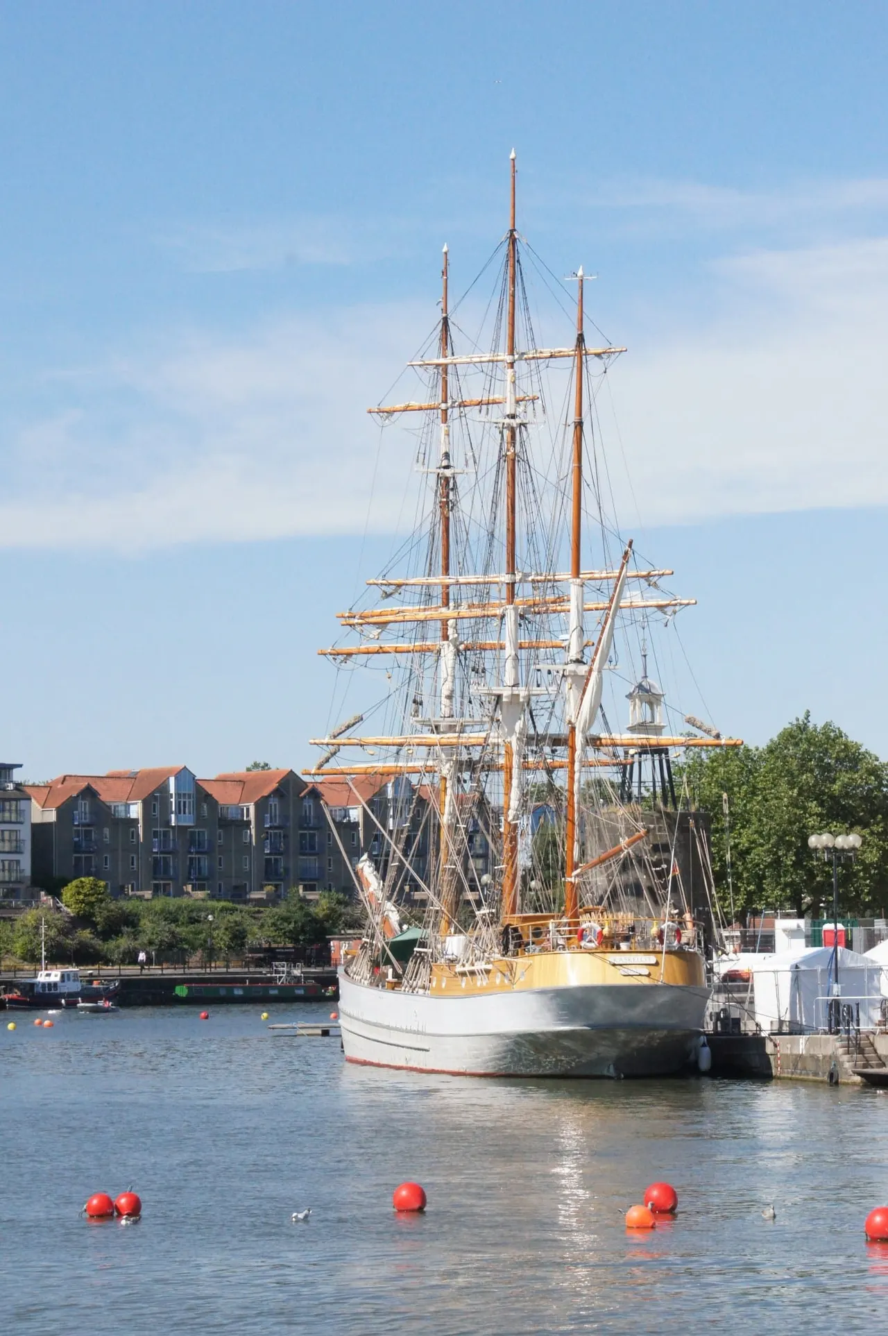 A tall ship with wooden masts and rigging moored at a riverside dock beside historic waterfront buildings.