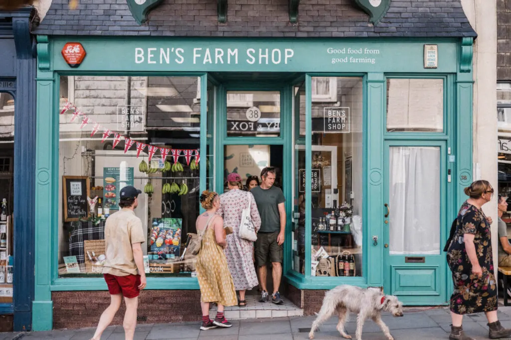 Customers enter a turquoise-fronted farm shop on a busy street as a dog walks past on the pavement.