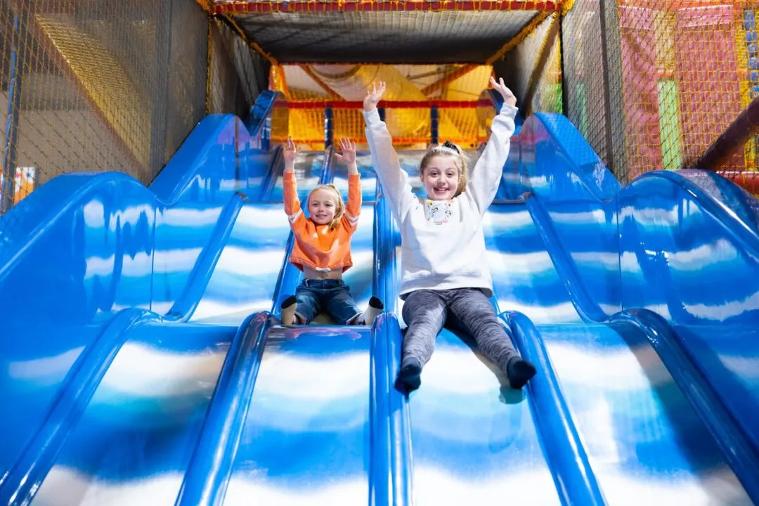 Two children sliding down a large blue indoor slide with arms raised, inside an inflatable play structure.