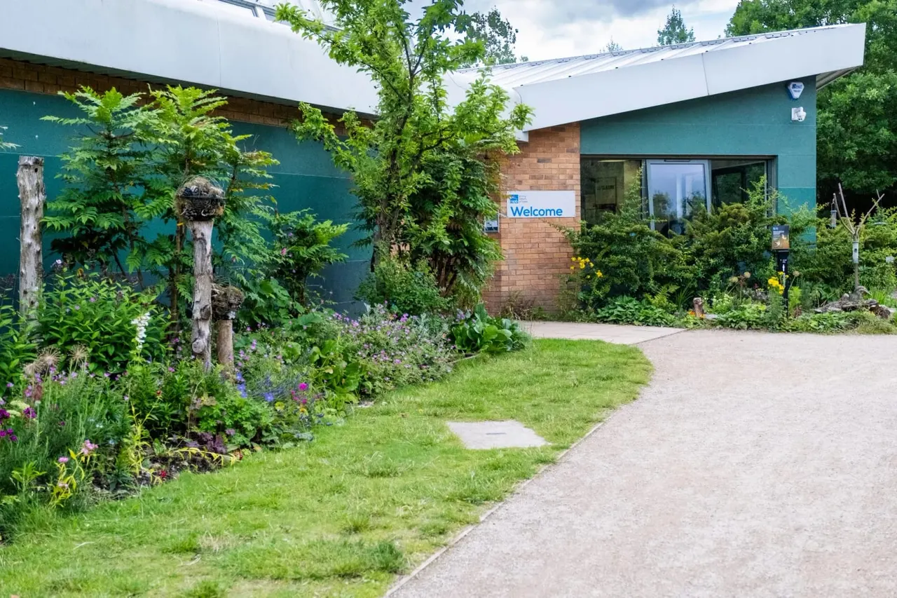 Pathway leading to a visitor centre with teal-painted walls, brick entrance, and surrounding gardens with flowering plants and green lawns.