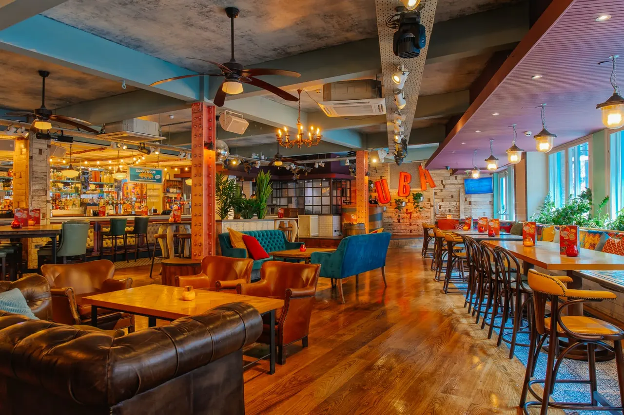 Spacious bar and lounge area with leather seating, wooden tables, colourful décor, and a bar counter visible in the background.