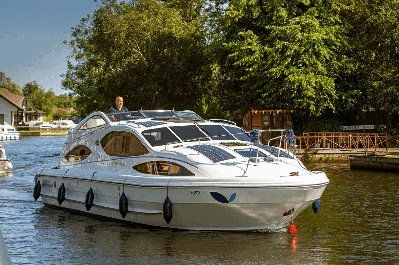 A white motor cruiser moored on a river with a person standing on the flybridge, surrounded by trees and riverside buildings.