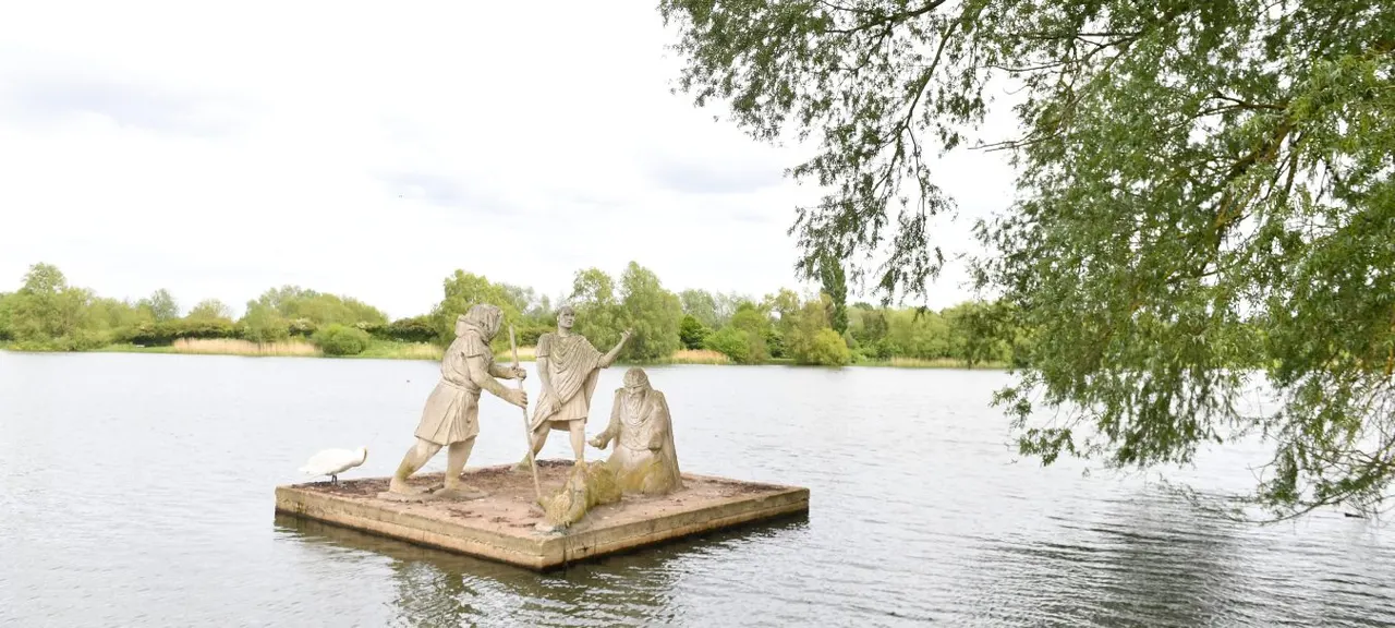 Stone sculptures of figures on a platform in a lake, with weeping willow branches framing the scene and wooded banks beyond.