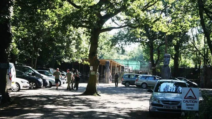 Shaded woodland car park with parked vehicles, visitors walking, and facilities buildings amongst mature trees.