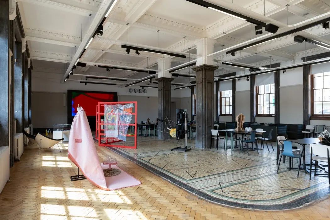 Spacious gallery interior with polished wooden floors, industrial ceiling, pink fabric installation, and red sculptural structure on display.