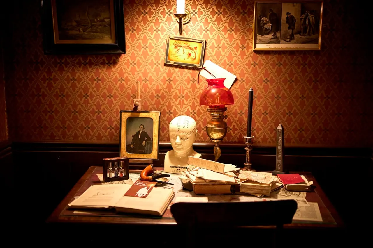 Museum display case with Victorian-era phrenology head, books, period lamps, portraits and documents against ornate patterned wallpaper.