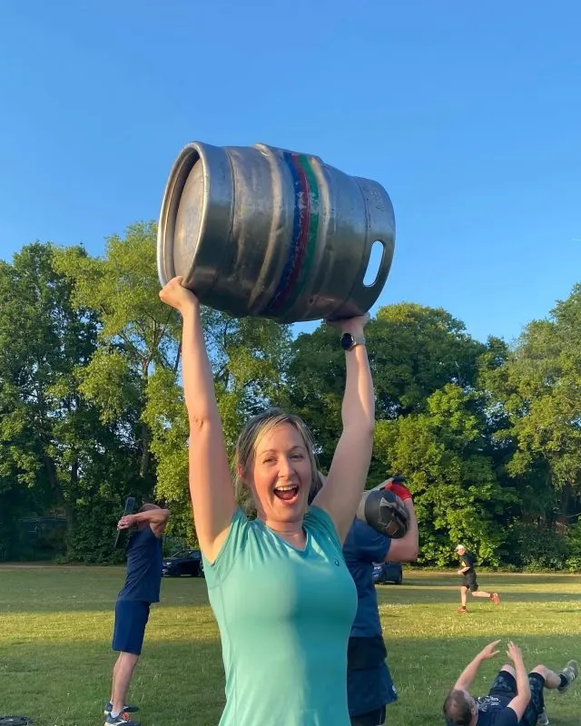 Woman holding a large barrel above her head during an outdoor fitness class in a park.