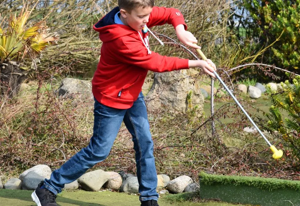 Child in red jacket playing miniature golf outdoors, swinging putter at ball on green course with rocks and vegetation.