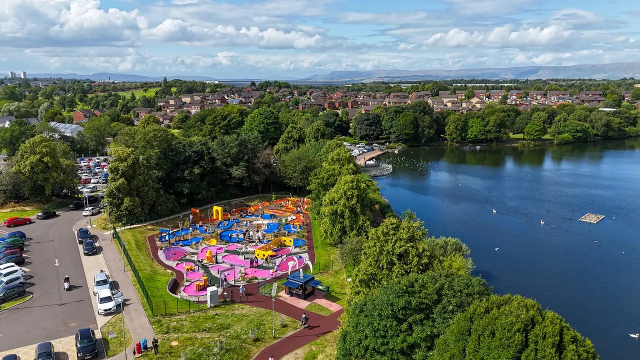 Aerial view of a children's adventure golf course with colourful pink and blue obstacles beside a river, surrounded by trees and residential suburb.