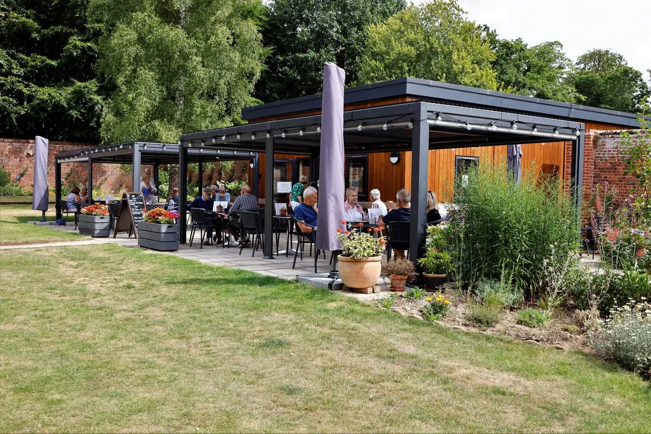 Visitors dining at outdoor covered seating areas beside a modern café building with garden planting and brick walls.