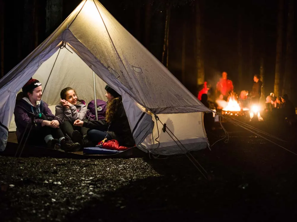 Three people sit inside an illuminated bell tent at night with a campfire and other campers visible in the dark forest background.