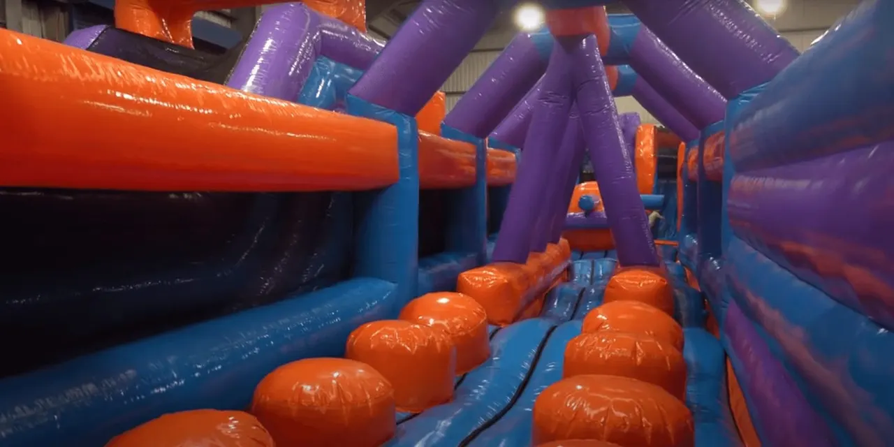 Interior of a large inflatable obstacle course with purple arch structures and orange inflatable bumpers lining a blue base path.