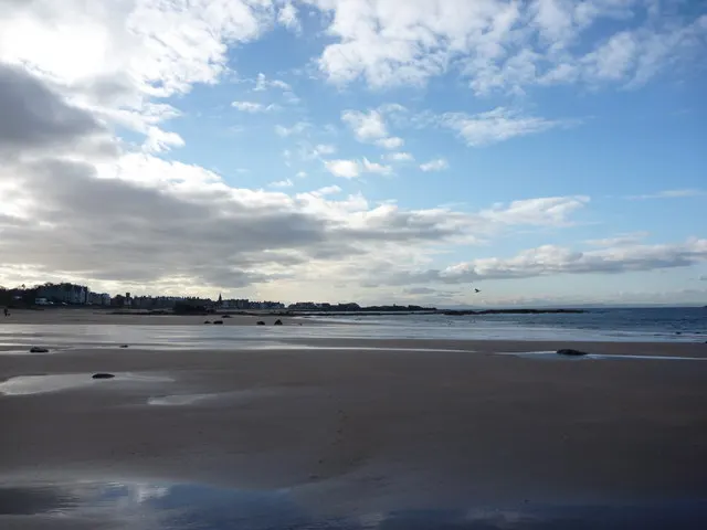 Expansive sandy beach at low tide with shallow water pools and coastal town visible across the bay under partly cloudy sky.