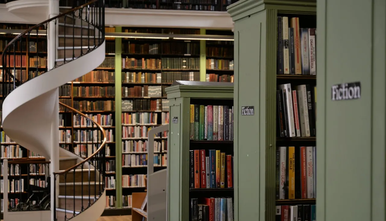 A spiral staircase beside bookshelves holding fiction collections in a modern library interior.