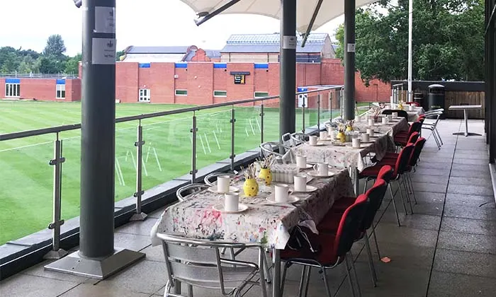A long table set for dining on an outdoor terrace overlooking a sports field and brick building.