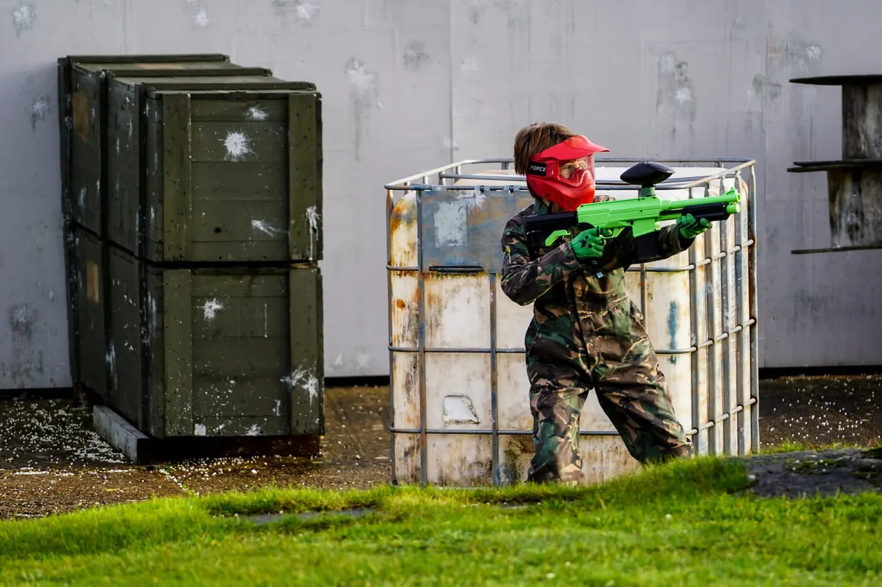 Player in red cap and mask aims bright green airsoft rifle behind weathered metal bunker during outdoor tactical game.