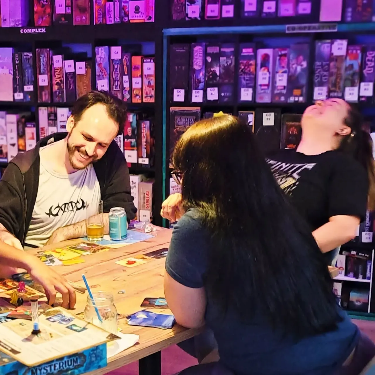 Three people playing board games at a wooden table in a brightly lit gaming venue with purple neon lighting and shelves of games behind them.